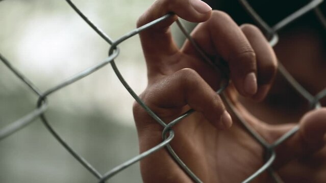 Close-up hand holding into metal fence barrier symbolizing entrapment behind cage. Person feeling imprisoned