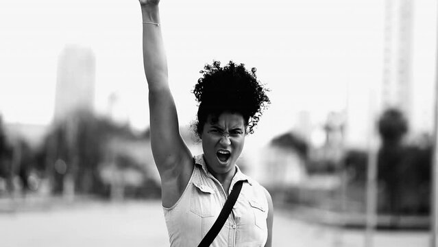 One determined young black woman raises fist in the air feeling empowered in dramatic black and white. African American female 20s person showing solidarity for cause
