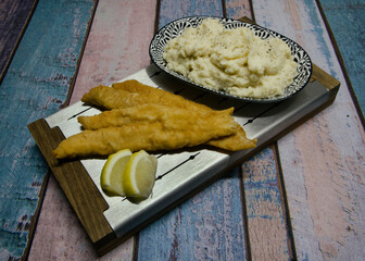 Closeup view of fried breaded fish filets with lemon and mashed potatoes in a metal board on the wooden table.	
