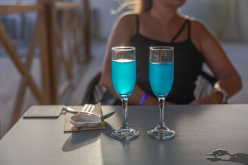 Close-up view of two glasses with blue alcoholic beverages and a girl sitting at a table in a restaurant with a blurred background. Curacao.