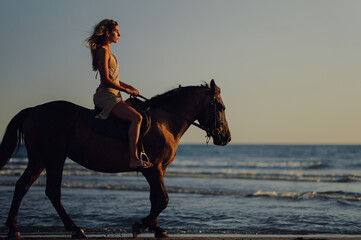 Side view of a woman enjoying horseback riding at sunset on a beach.