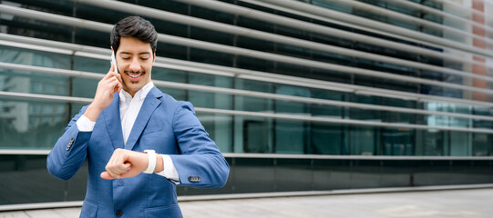 A young Hispanic businessman in a blue suit confidently checks the time on his wristwatch while making a phone call, against a backdrop of modern corporate architecture. Concept of time management