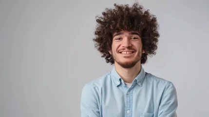 Fotobehang cheerful man with curly hair, smiling at the camera, arms crossed, wearing a light blue casual shirt, standing against a light gray background. © MP Studio