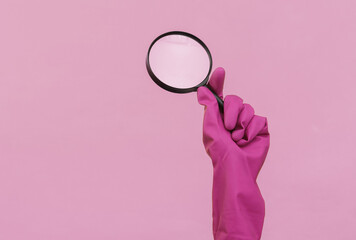 Hand in purple rubber cleaning glove holding magnifying glass on pink background. House cleaning and housekeeping concept - obrazy, fototapety, plakaty