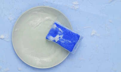 Sponge and plate with foam on a blue background. Washing dishes concept. Top view