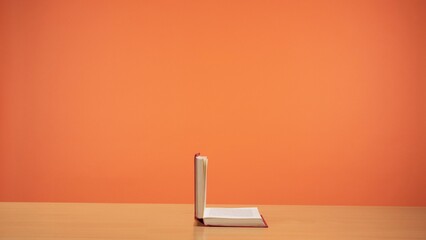An old open book with red cover on a wooden table on a bright orange background. - obrazy, fototapety, plakaty