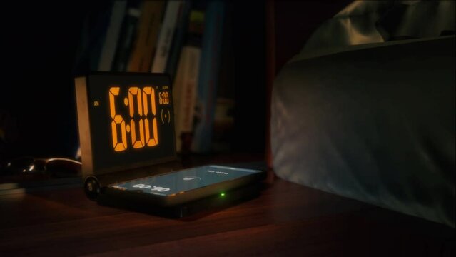 Digital alarm clock with phone charger and orange clockface is waking up at 6 AM. The numbers on the clock screen changes from 5:59 no 6:00 AM. And the alarm goes off on the phone that is charging.
