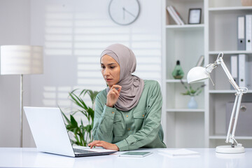 A dedicated professional wearing a hijab is deeply focused as she works on her laptop at a bright...