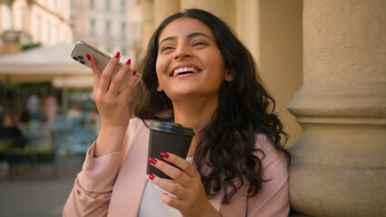 Young smiling happy businesswoman Indian Arabian ethnic woman girl female business worker listening voice message smartphone drink cup coffee receiving audio using cellphone social media outside town