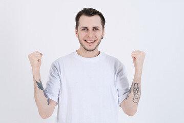 Attractive caucasian young man with tattoos in casual clothes looking at camera, smiling and showing winner gesture isolated on white studio background