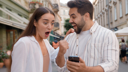 Caucasian happy couple enjoy excited man woman shocked lucky success victory mobile phone smartphone reading message good news amazed surprised open mouth city outdoors urban gen z win wonder awesome