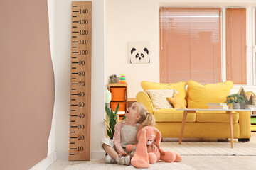 Cute little girl with toy near wooden stadiometer at home