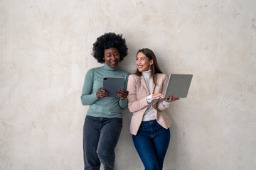 Two multiethnic businesswomen using digital tablet and laptop while standing against a studio...