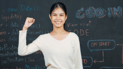 Young student celebrate her successful plan while raise her arm. Happy teenager looking at camera while standing at blackboard with engineering code and prompt written in STEM class. Edification.
