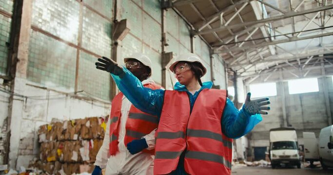 A brunette girl in safety glasses and a special uniform in an orange vest and a white helmet walks with her employee a man with Black skin through a huge waste paper processing plant