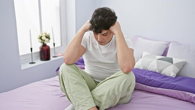 A distressed young man sitting on a bed in a bedroom, holding his head in a worried manner.