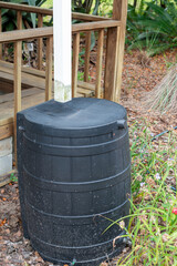 A black plastic rain barrel with a white metal downspout connected to the lid of the container. The...