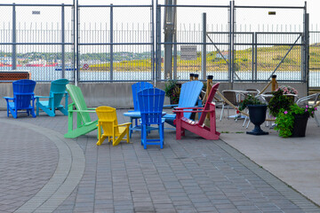A row of colorful recycled wood Adirondack chairs on a sidewalk next to a large red brick building...