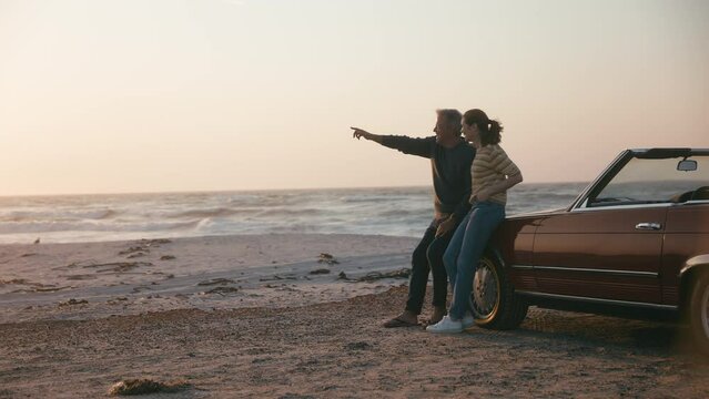 Retired senior couple on vacation standing by classic convertible sports car at beach watching morning sunrise on road trip  - shot in slow motion - Powered by Adobe