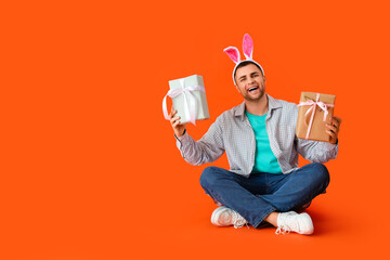 Happy young man in bunny ears with gift boxes sitting on orange background. Easter celebration