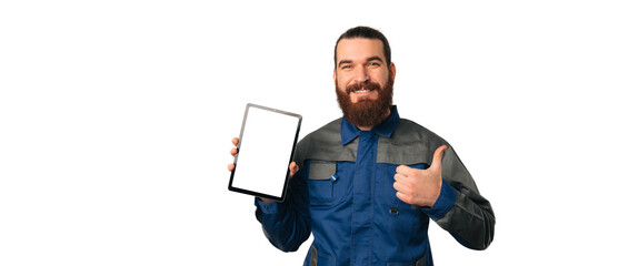 Young repairman wearing a blue uniform is holding a tablet with blank screen an d athumb up. Banner size shot.