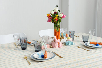 Beautiful table setting for Easter decorated with bouquet of carrots and flowers against white wall