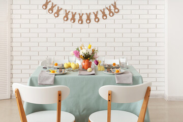 Beautiful table setting for Easter decorated with bouquet of carrots and flowers near white brick wall