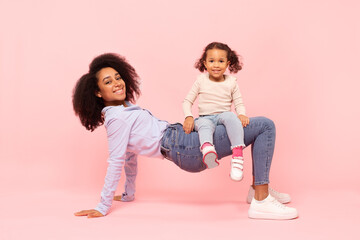 Joyful little black daughter having fun with young mother, girl sitting on mom's legs like on chair against pink studio background