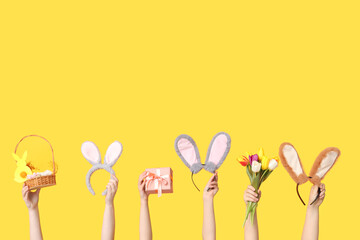 Female hands holding Easter bunny ears headbands with basket, gift box and tulips on yellow background