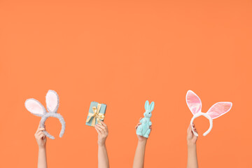 Female hands holding Easter bunny ears headbands with gift box and toy rabbit on orange background