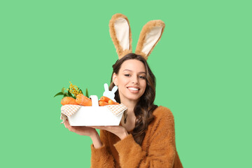 Happy young woman in Easter bunny ears headband holding basket with carrots on green background