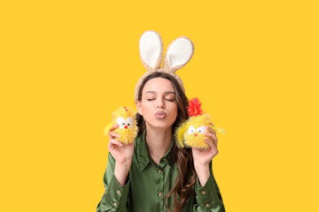 Young woman in Easter bunny ears headband with toy chickens blowing kiss on yellow background