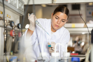 Focused female chemist pipetting blue reagent into solution in test tube while performing...