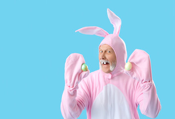 Senior man in Easter bunny costume with painted eggs on blue background