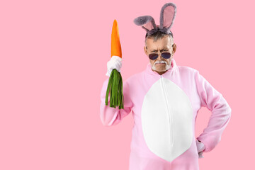 Cool senior man in Easter bunny costume with plush carrot on pink background
