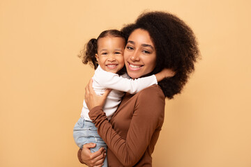 Portrait of cute black girl and her mother hugging and smiling at camera, beige background, healthy united family, mother daughter bonding concept