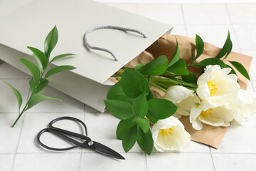 Paper shopping bag with tulips, green twigs and scissors on white tile background