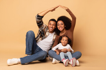 Fototapeta na wymiar Young black family oh three sitting on floor and making roof shape with their hands above, symbolizing home, beige studio background