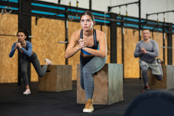 Concentrated young girl in fitness attire performing Bulgarian split squats using wooden plyometric box at gym. Bodyweight training concept.. - obrazy, fototapety, plakaty