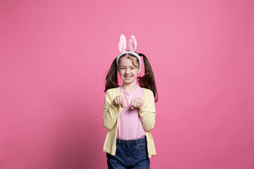 Joyful young girl jumping around like a rabbit in front of camera, wearing bunny ears and celebrating easter sunday holiday. Smiling little toddler laughing and having fun over pink backdrop.