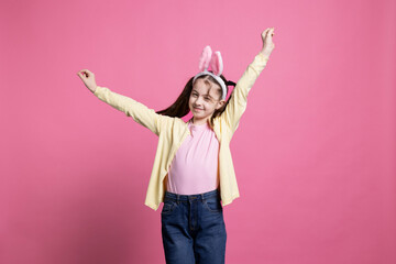 Sweet small child dancing in front of studio camera, posing with confidence and having fun. Little girl acting carefree with dance moves over pink background, wears bunny ears.