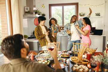 Cheerful young group of diverse friends holding drinks and dancing at home kitchen during party time