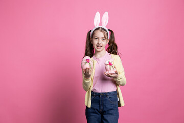 Small girl with bunny ears holding pink easter decorations in front of camera, presenting her...
