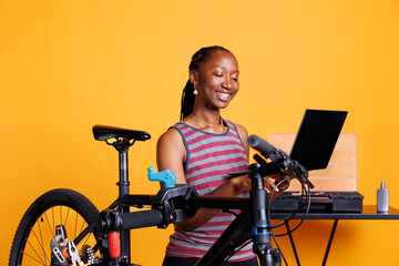 Energetic black woman inspecting and fixing bike, utilizing laptop and specialized toolkit for...
