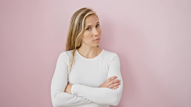 Skeptic young blonde damning with a nervous and disapproving facial expression, standing with arms crossed over isolated pink background