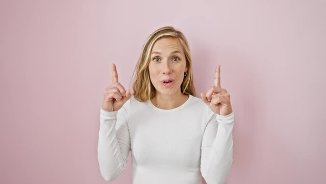 Young, beautiful blonde woman overjoyed with girlish joy - pointing upwards with a look of surprise on her face, standing against a whimsical isolated pink backdrop.