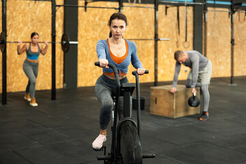 Fototapeta na wymiar Woman performs exercises with stationary bicycle. Female gym visitor performs series of repetitions of movements on simulator. Shot of muscular man in sportswear and good physique on grey background