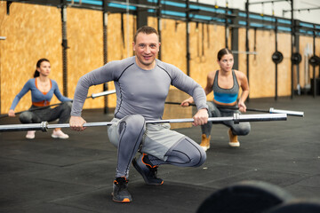Smiling athletic man squatting with empty barbell bar in hands, preparing to perform exercises...