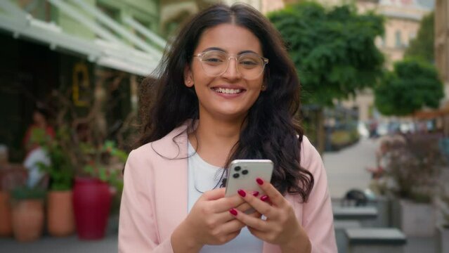 Happy successful smiling woman Indian Arabian ethnic female girl customer user holding mobile phone outside city street cafe. Positive client nodding head yes good reaction approve technology gadget
