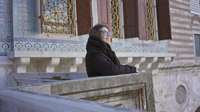 A smiling woman in glasses leaning on an ornate stone balustrade at topkapi palace, istanbul, exudes a tourist vibe amidst ottoman architecture.